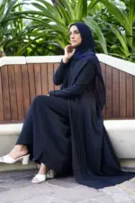 Woman wearing a black pleated blazer set with a matching hijab and white heeled sandals, seated on a wooden bench surrounded by lush green plants. The outfit features elegant pleats and a modest, flowing silhouette, styled for a formal or semi-formal look